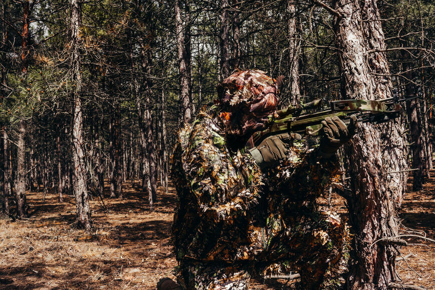 camouflaged sniper hunter with tactical crossbow sitting in woods